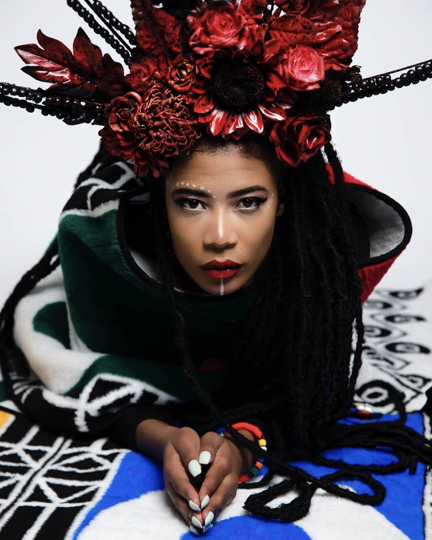 A woman crowned with red roses, gaze direct, resting on the Unity Throw — the Idzila geometric motif beneath her. Editorial portrait of the brand's anchor piece.